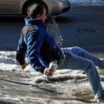 Man in jeans and blue winter pullover holding a beverage slipping on the ice. Orange hazard cone and lower half of a silver car in the background.