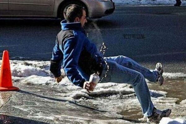 Man in jeans and blue winter pullover holding a beverage slipping on the ice. Orange hazard cone and lower half of a silver car in the background.