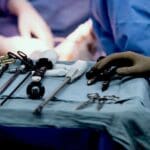 Close-up of surgical instruments on a tray. Blurred image of two surgeons' gloved hands at work in the background.