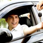 New male teen driver in white shortsleeved polo shirt sits behind the wheel of a car. Adult hand is handing him a car key through the window.