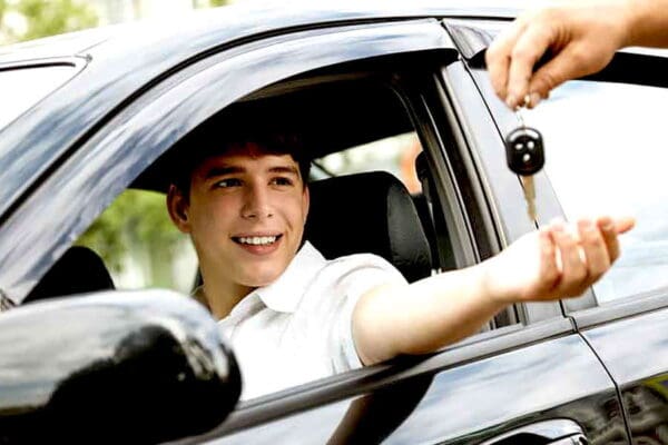 New male teen driver in white shortsleeved polo shirt sits behind the wheel of a car. Adult hand is handing him a car key through the window.