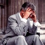 Grey haired many in pale grey suit and white buttondown sits on the steps of a courthouse with his head in his hands.