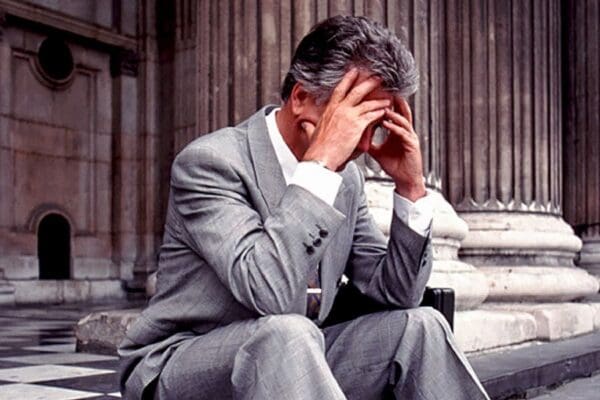 Grey haired many in pale grey suit and white buttondown sits on the steps of a courthouse with his head in his hands.